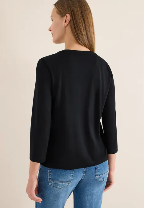 Frau von hinten in schwarzem 3/4-Arm-Oberteil und blauen Jeans vor neutralem Hintergrund